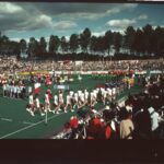 colour photo of a hockey field with groups of people in processions carrying names of countries they are from: france and pakistan can be read. there are spectators all around the field.