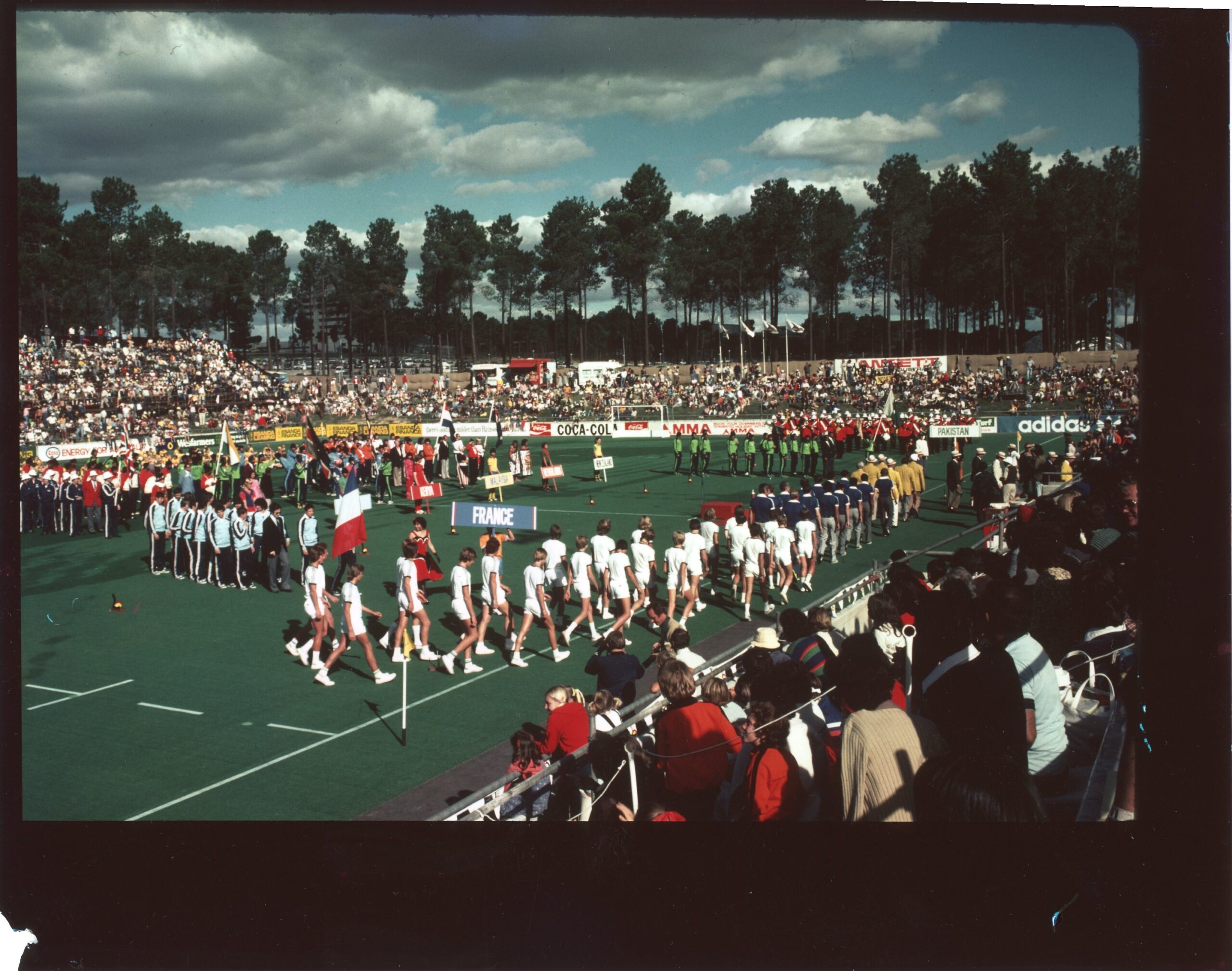 colour photo of a hockey field with groups of people in processions carrying names of countries they are from: france and pakistan can be read. there are spectators all around the field.