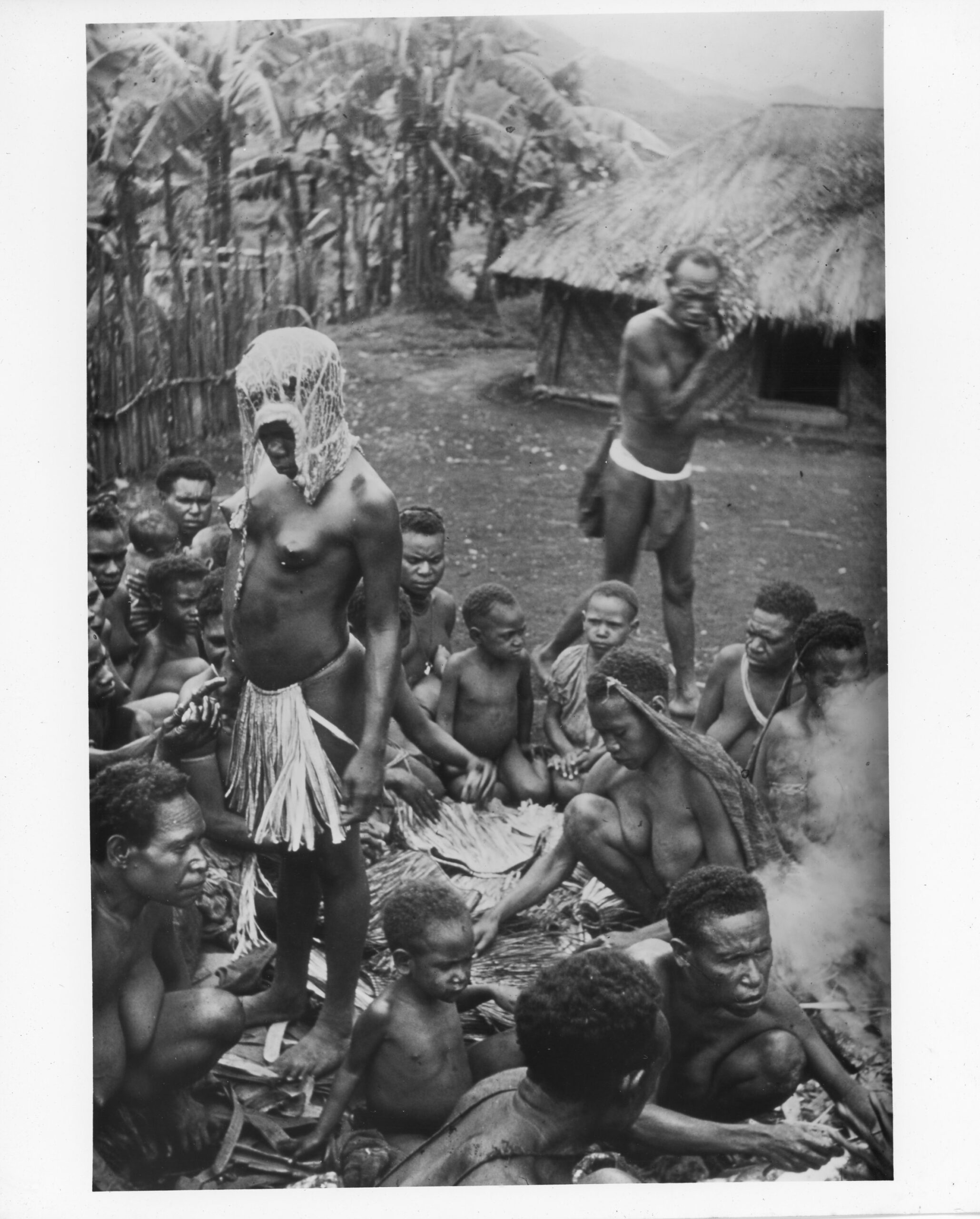 black and white photo of a Melanesian ceremony, a group of people sit on the ground around a bride wearing a pig's stomach on her head and a grass skirt.