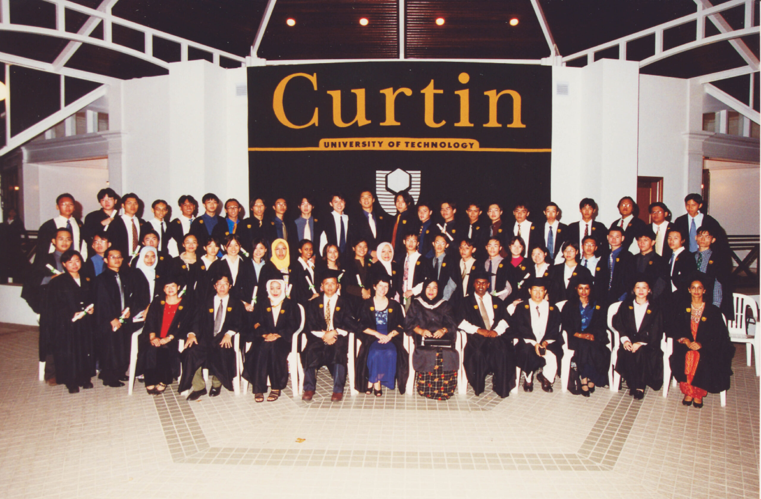 a formal portrait of three rows of student graduates wearing black academic gowns and coloured sashes, holding their graduation certificates. behind them is a large black sign with 'Curtin University of Technology' in yellow.