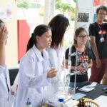 students in white lab coats stand at a stall speaking to attendants about their project at the innovation expo. on the table is chemistry beakers and equipment.
