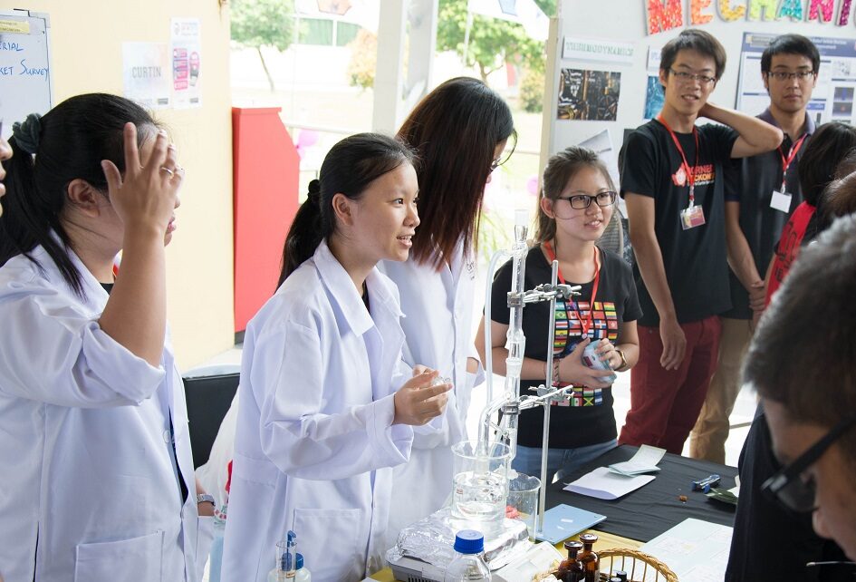 students in white lab coats stand at a stall speaking to attendants about their project at the innovation expo. on the table is chemistry beakers and equipment.