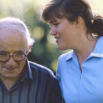 A nursing student with brown hair wearing a light blue nursing uniform holds the arm of an elderly man as they walk.