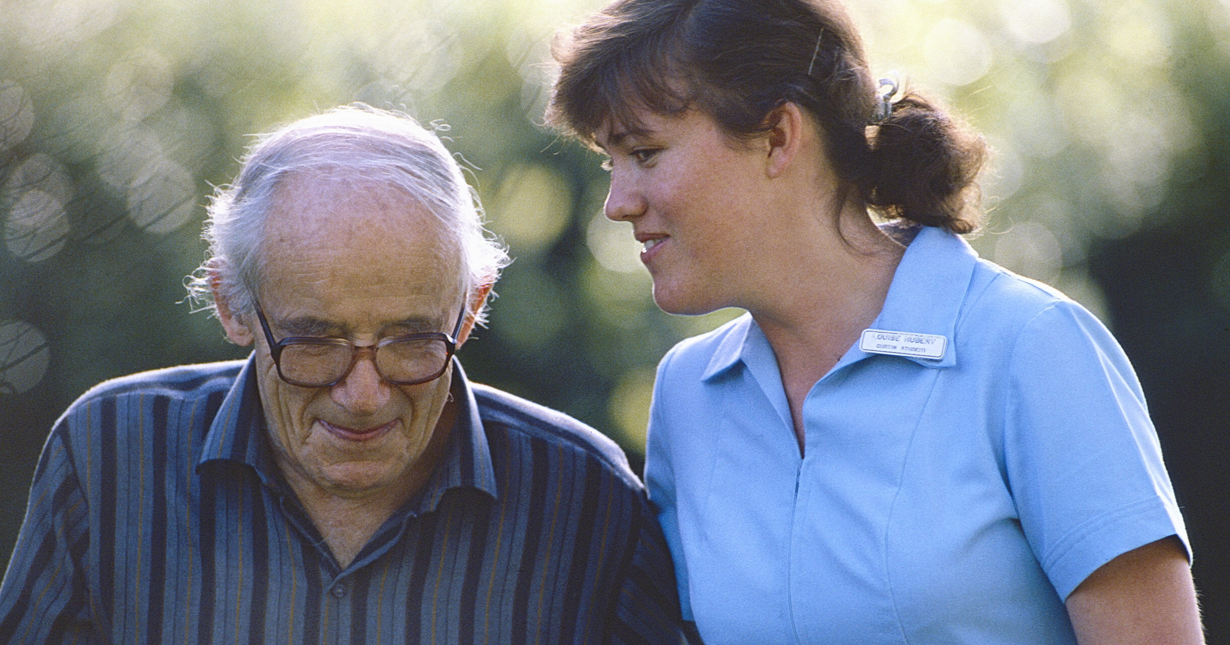 A nursing student with brown hair wearing a light blue nursing uniform holds the arm of an elderly man as they walk.