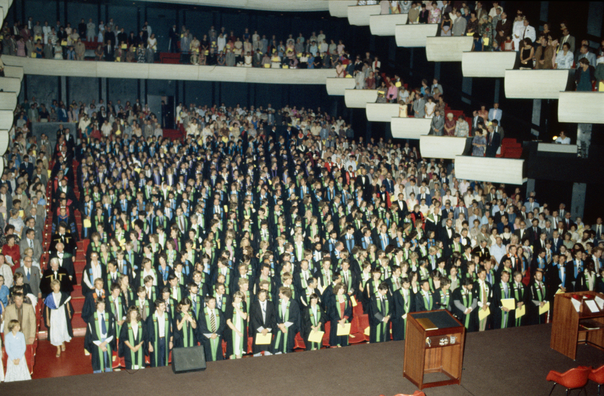 a concert hall room at a graduation ceremony. the seats are filled with graduates wearing black academic gowns and coloured sashes, and other attendees in plain clothing.