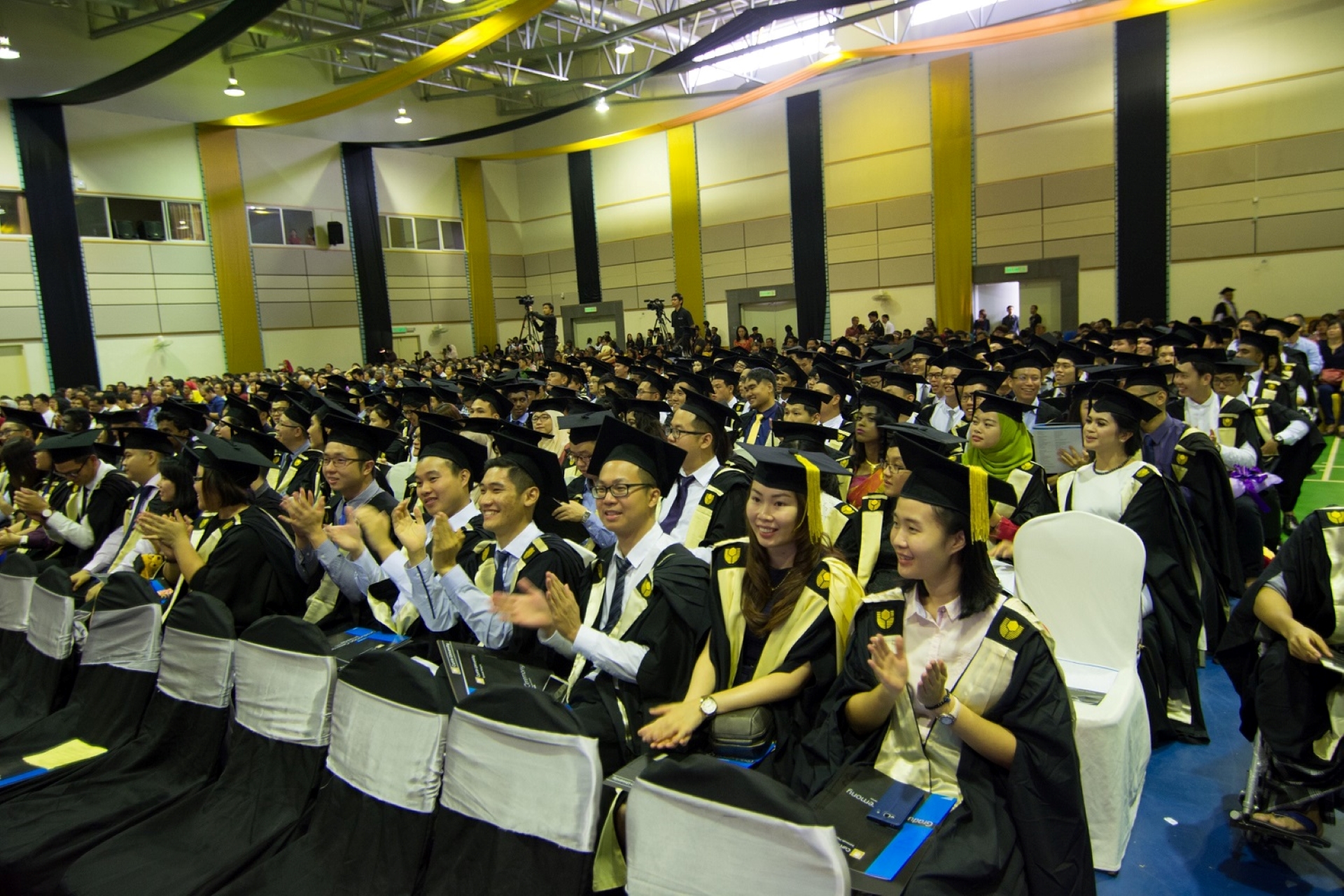 A room full of Curtin Singapore students sitting wearing academic gowns, sashes and mortarboards, appauding at their graduation.