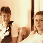 Sepia brown tone image. A woman with short hair and large glasses and badge is looking at a young woman, who is looking at the camera.