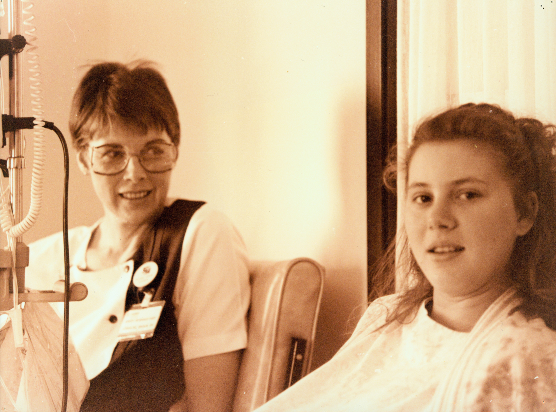 Sepia brown tone image. A woman with short hair and large glasses and badge is looking at a young woman, who is looking at the camera.