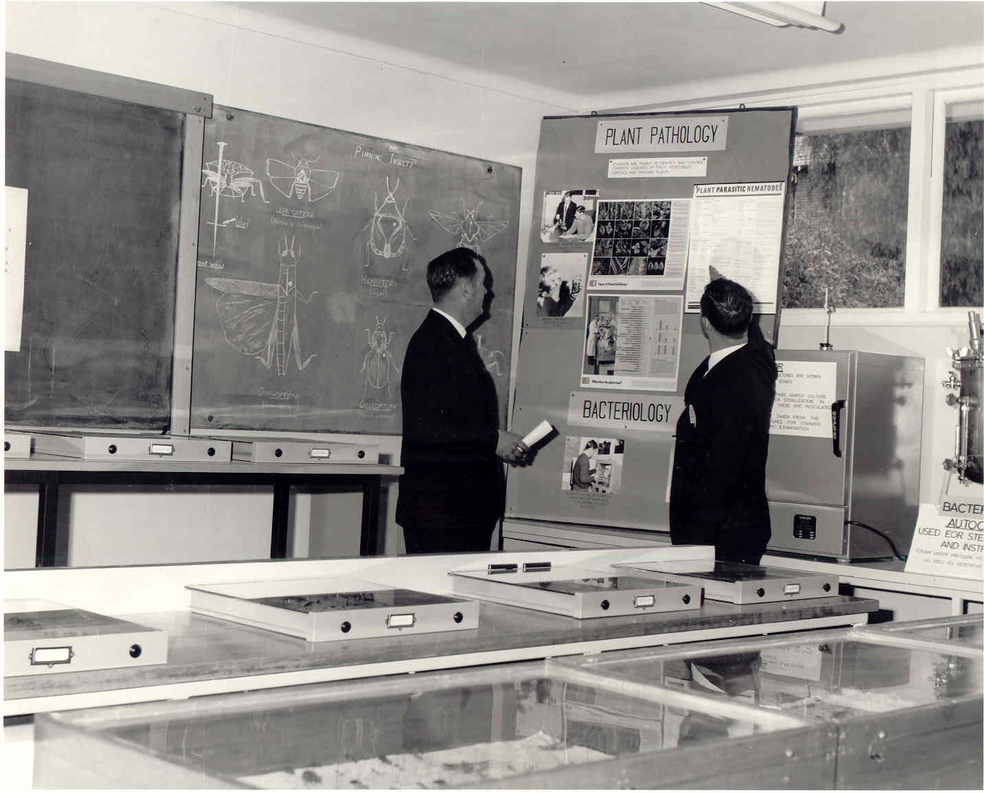 Black and white photo of two people in a classroom, pointing to a blackboard that reads "plant pathology"