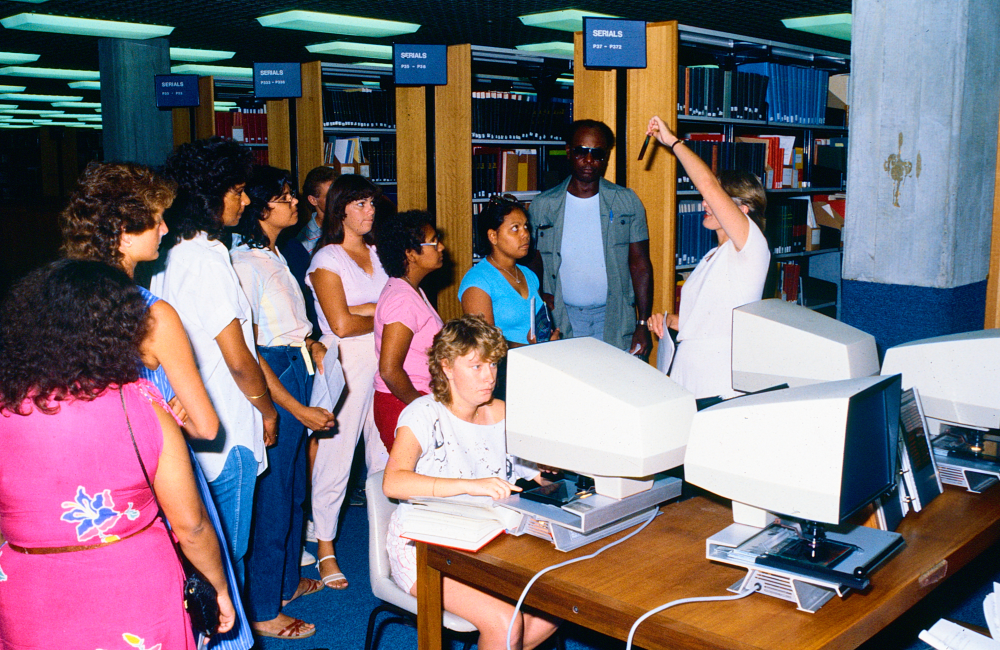 a student sits at a large white computer, behind her a staff member is showing a group around. library book shelves are in the background.