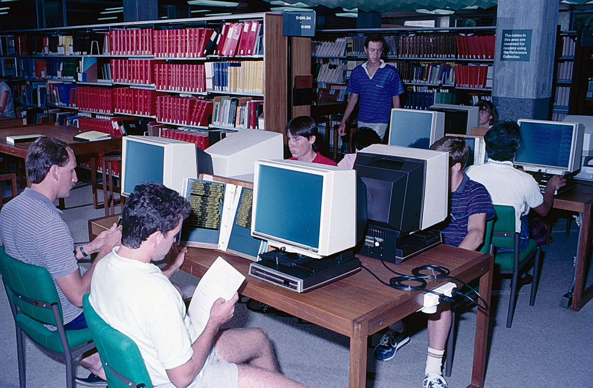 students at catalogue machines, library bookshelves in the background.