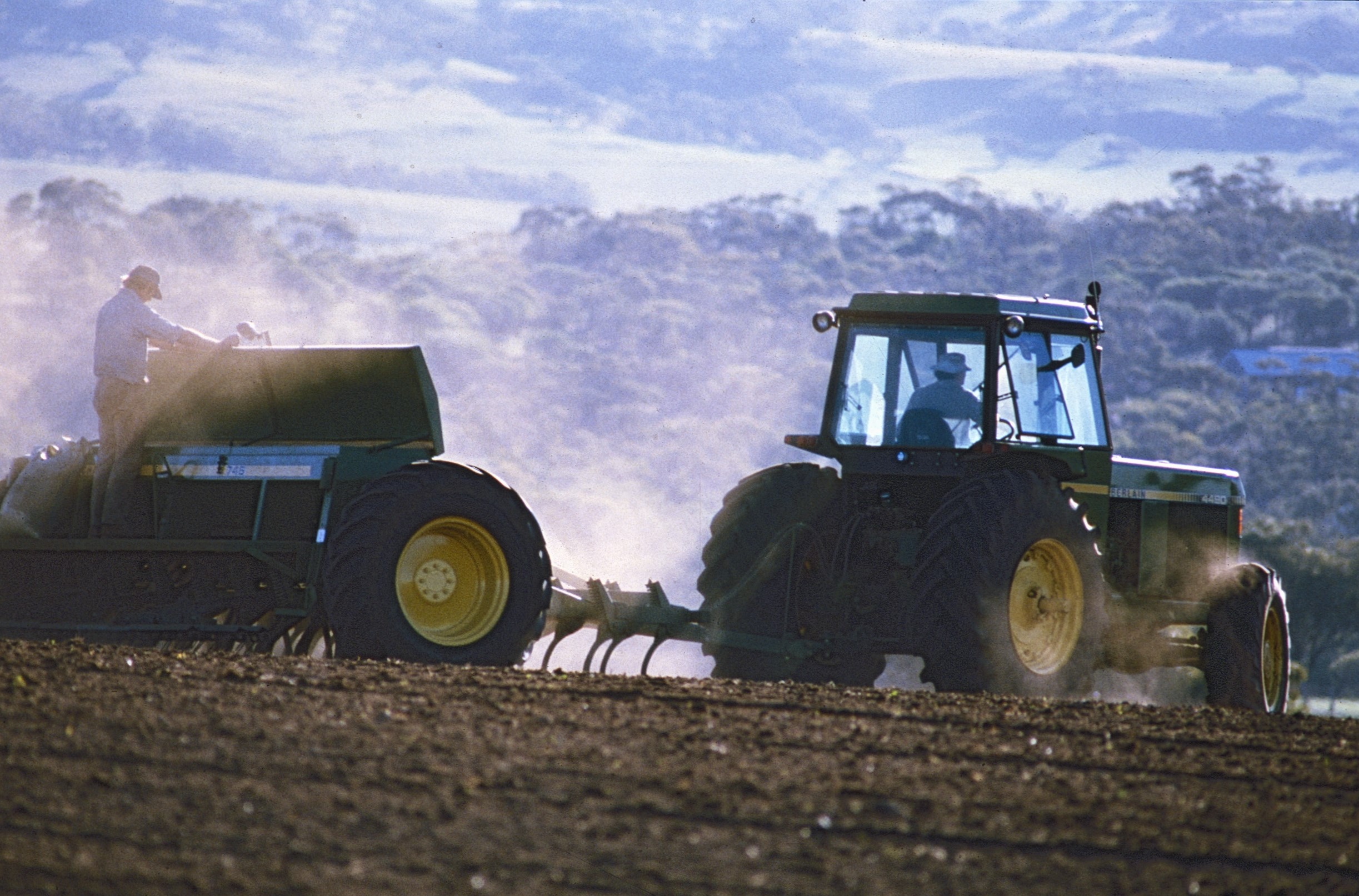 A tractor is pulling a trailer over a crop field, creating dust in the background.