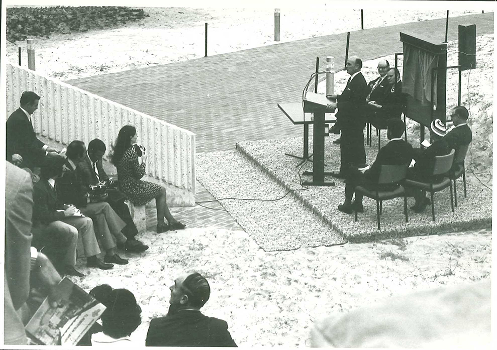 black and white photo of a man in a suit speaks into a microphone. people are sitting behind him and to the left an audience sits in Atkinson Forum amphitheatre.