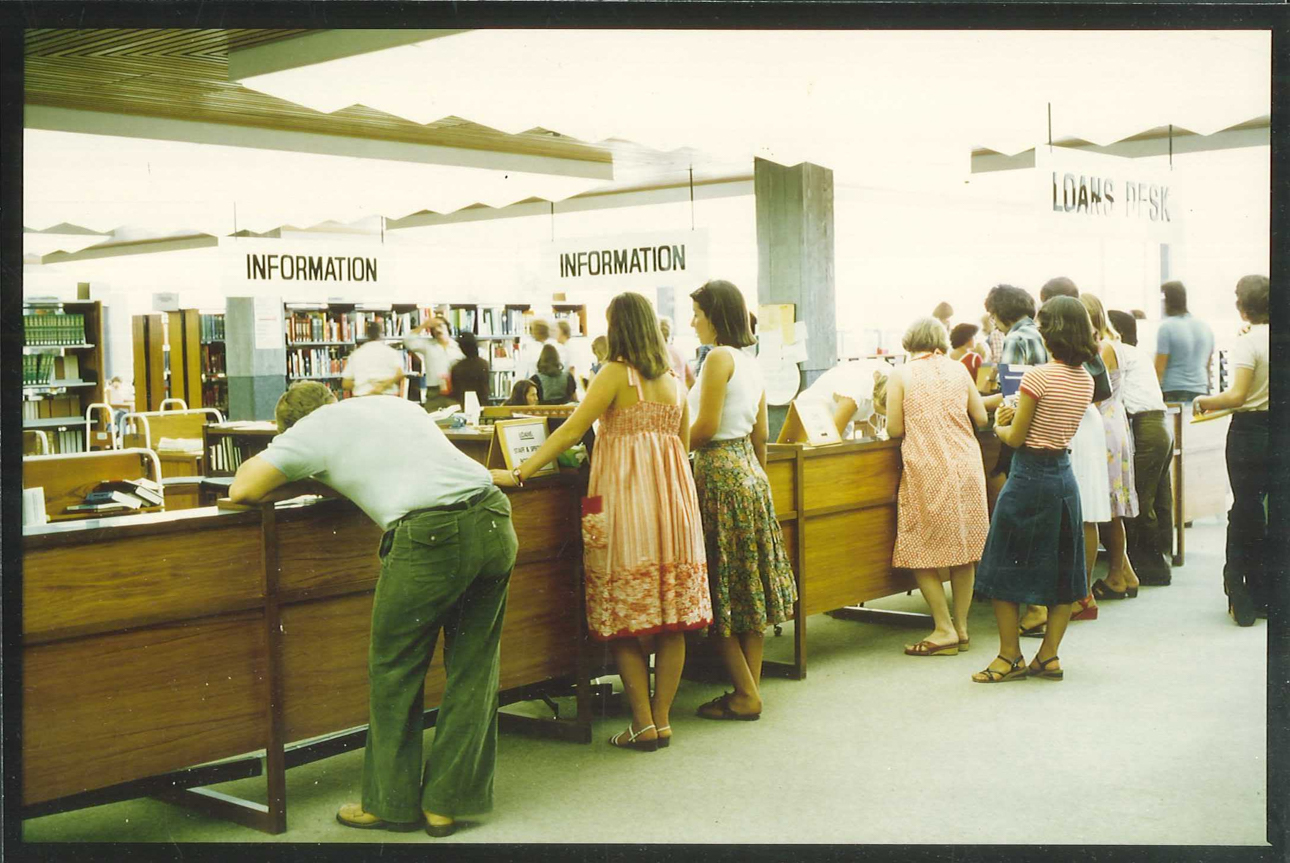 faded colour photo of people waiting at a wooden desk in the library