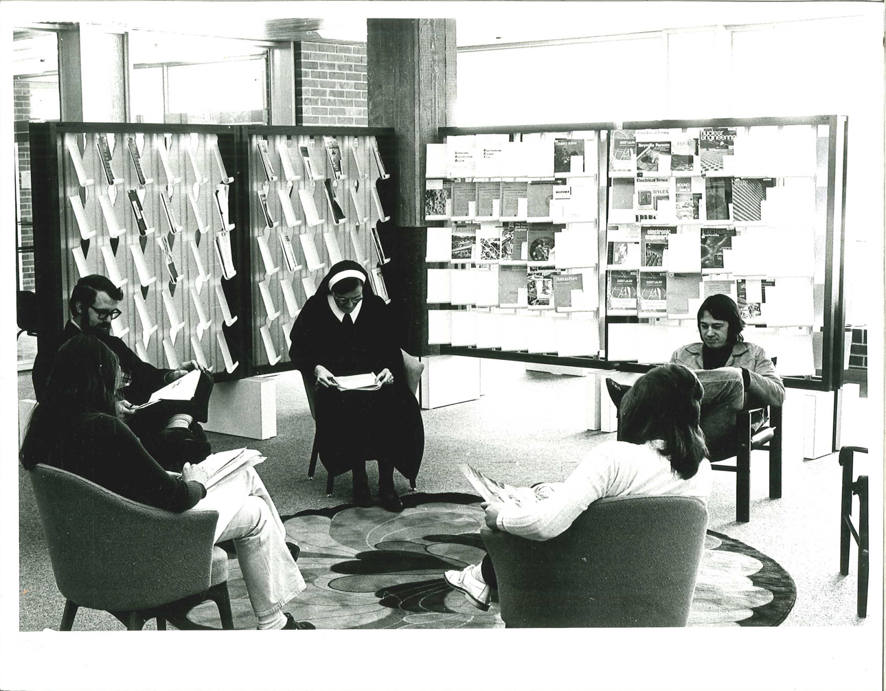 black and white photo of five people sitting in lounge chairs reading in the library