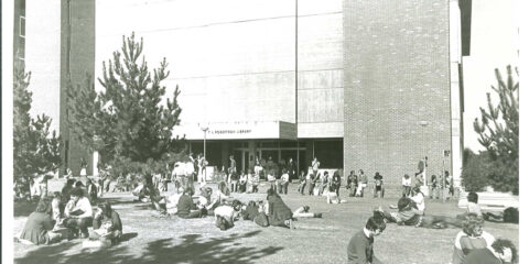 black and white photo of students sitting on the grass in front of the concrete library building.