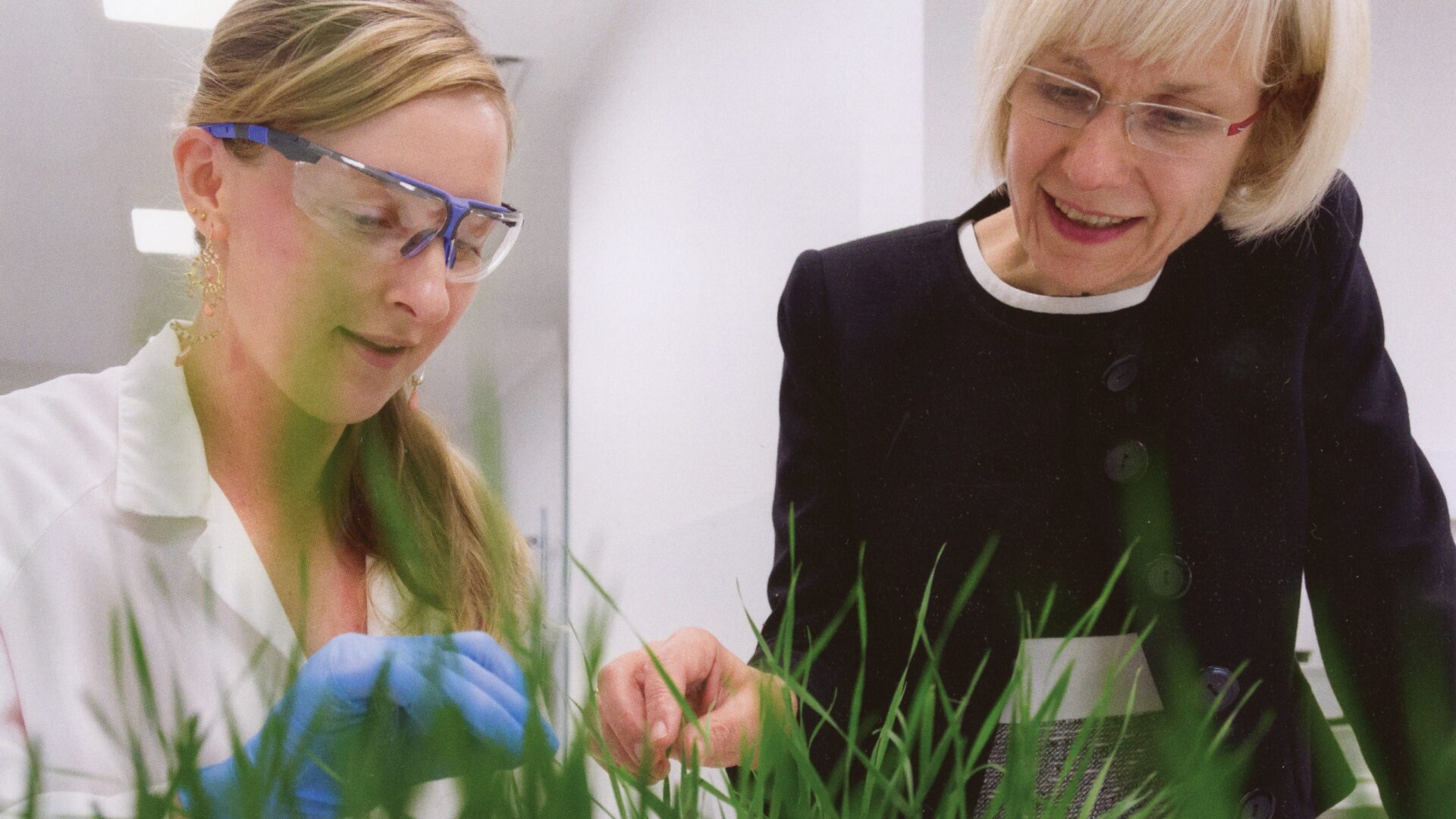 A person in a white lab coat studies some plant samples up close, next to her Deborah Terry looks over and is smiling. She has short blond hair and glasses.