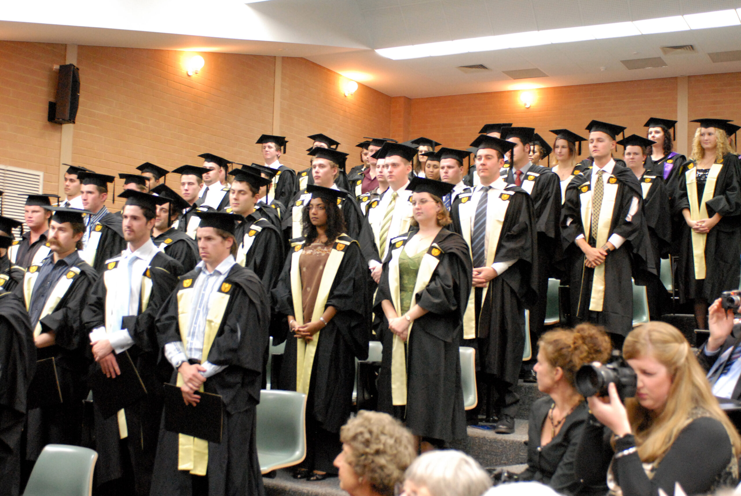 A lecture theatre full of students standing in black academic gowns and wearing gold sashes and mortarboards.